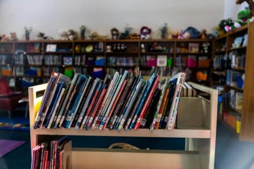 A warm library interior featuring a book cart in focus with colorful children's books on shelves.