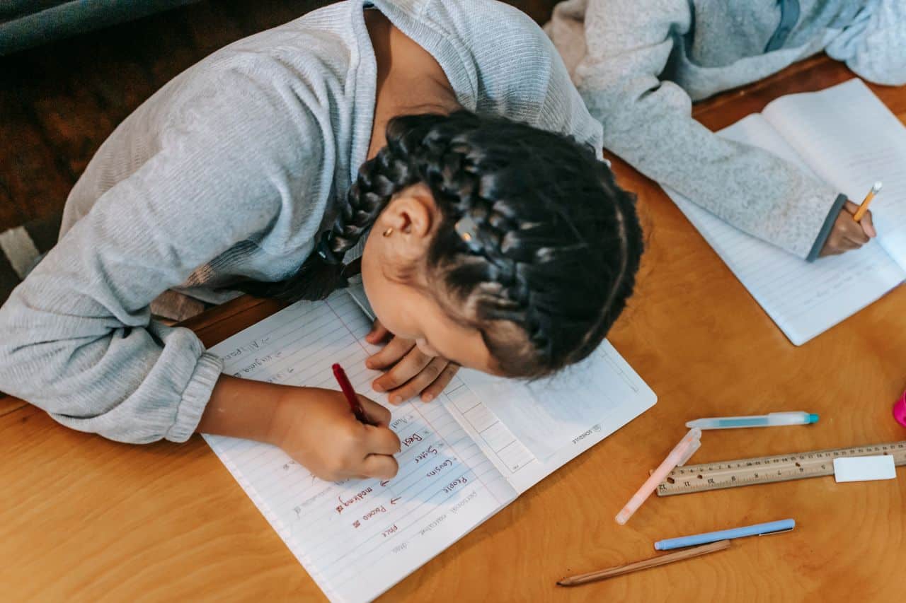 From above of focused kids writing in copybooks while doing homework together at wooden table